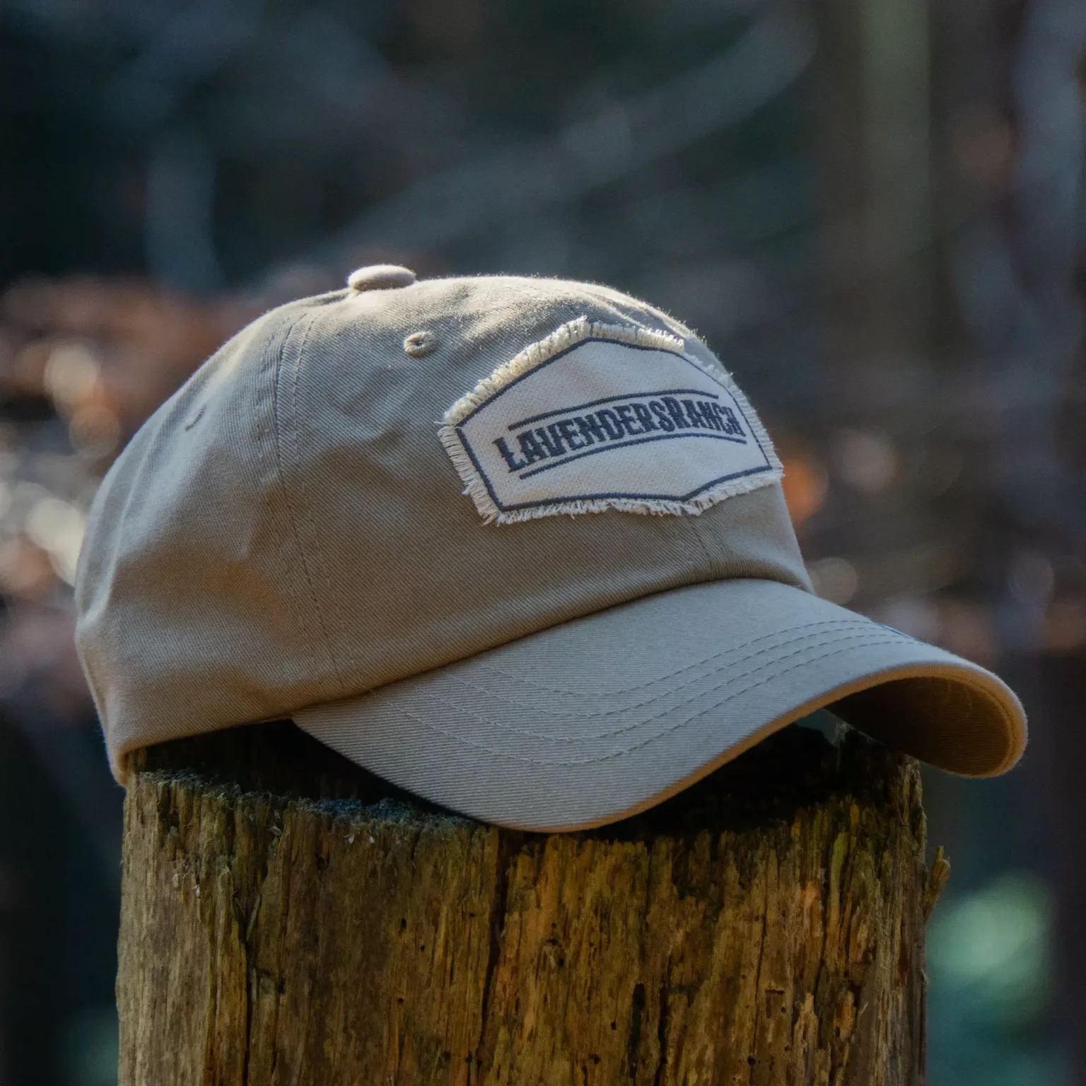 LavendersRanch Badge Cap light khaki – close-up of embroidered patch cap resting on a rustic wooden post in forest light