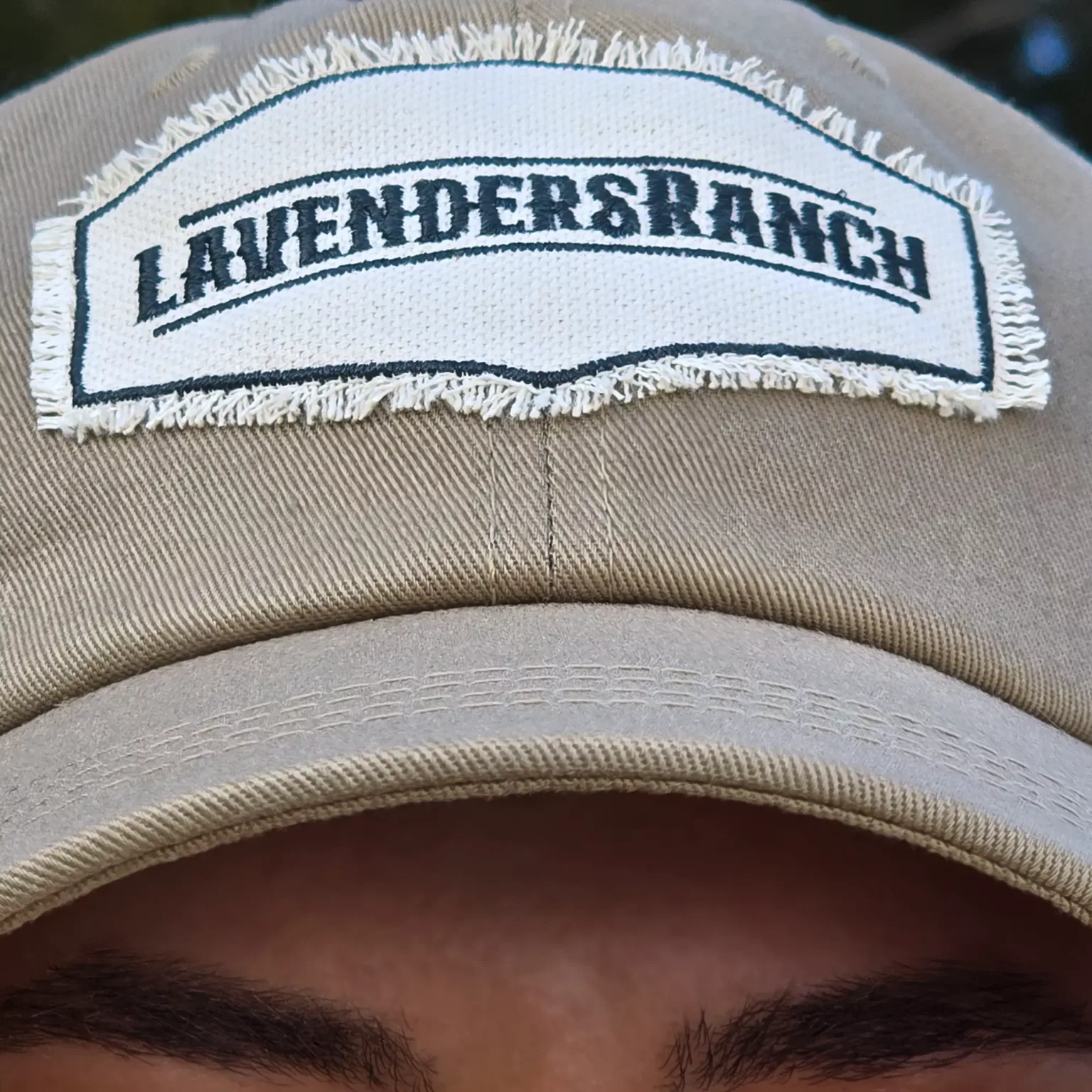 LavendersRanch Badge Cap light khaki – extreme close-up of frayed embroidered logo patch on cap above model’s eyes
