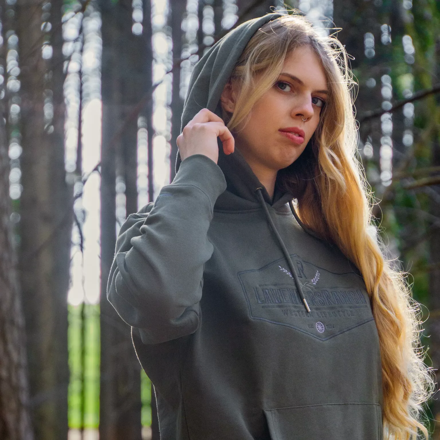 LavendersRanch Hoodie khaki green worn by woman in forest setting, showcasing embroidered chest logo and raised hood.