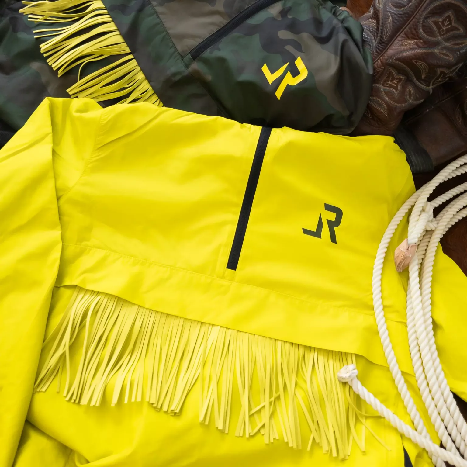 LavendersRanch Windbreaker Jacket camouflage – flat lay of camo jacket with yellow fringe and logo, next to citrus version, cowboy boots, and a rope lasso.
