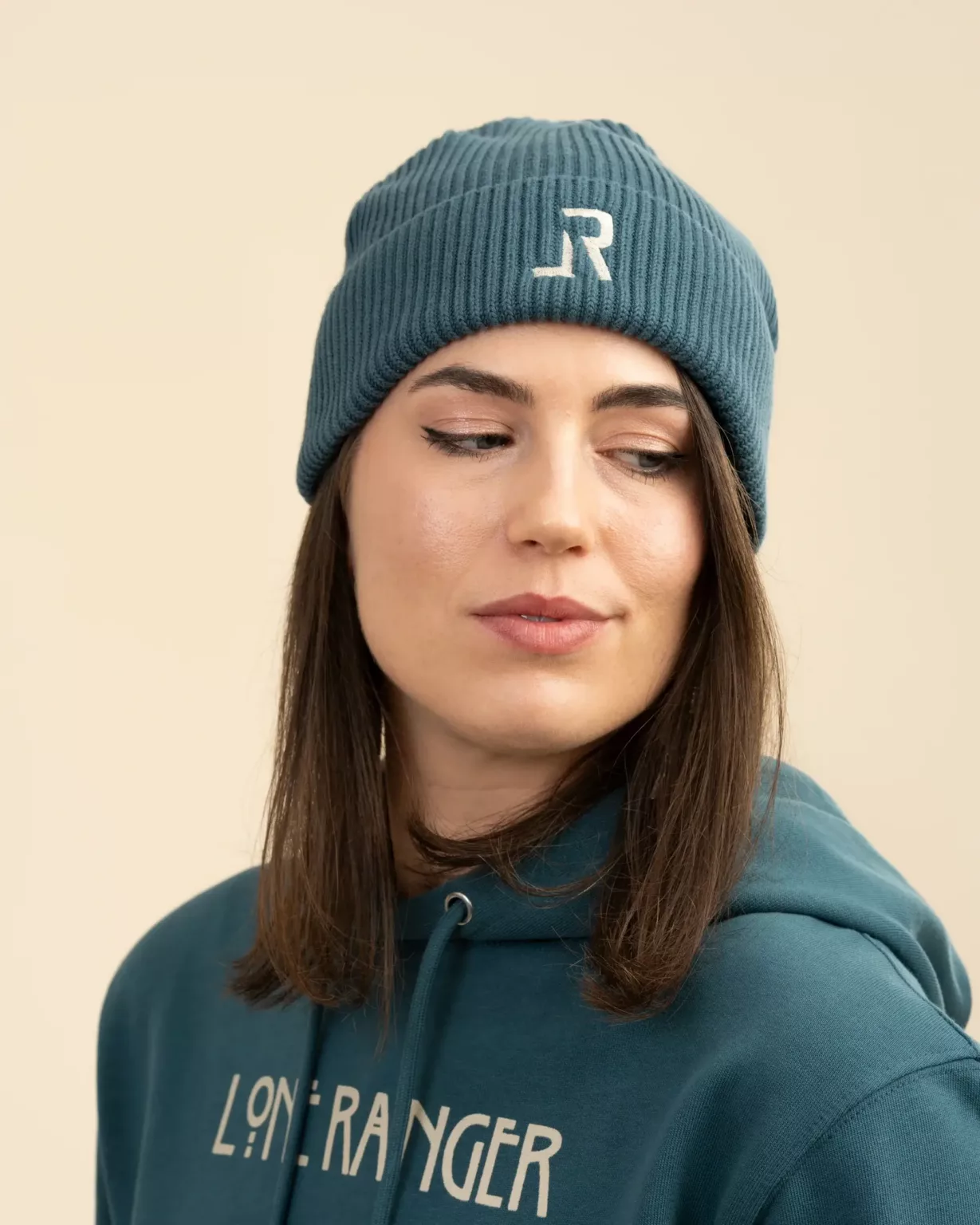 Close-up of a woman wearing a teal-blue-colored "LR" beanie with a "Lone Ranger" hoodie, showcasing sustainable fashion by LavendersRanch (LR).